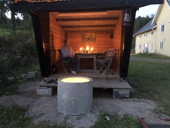 Una cabaña de madera con una mesa y sillas, un hoyo de fuego y una persona sentada junto al fuego.