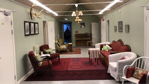 Ein geräumiges Wohnzimmer mit hoher Decke, Holzbalken und einem roten Teppich mit Muster. Mehrere Sessel und ein Sofa laden zum Verweilen ein, im Hintergrund steht ein Klavier.