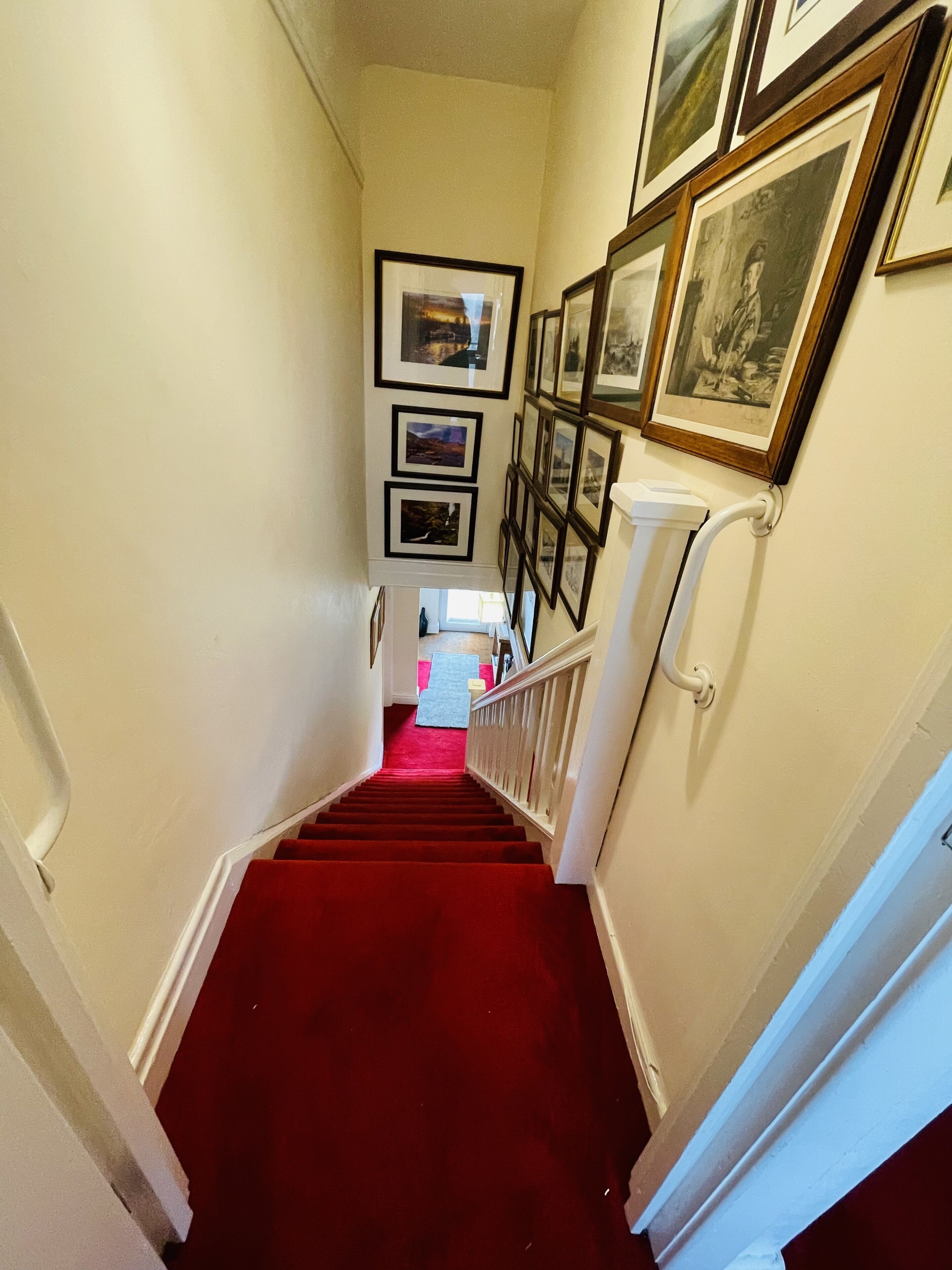 Un escalier recouvert d’une moquette rouge menant à un palier orné de tableaux encadrés.
