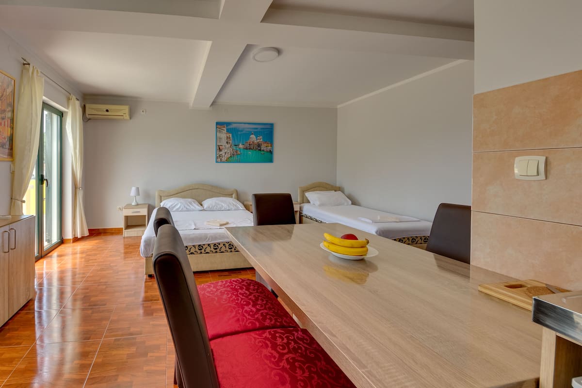 A hotel room with a wooden dining table, a red cushioned bench, two single beds, a painting of Venice, and a window with curtains.