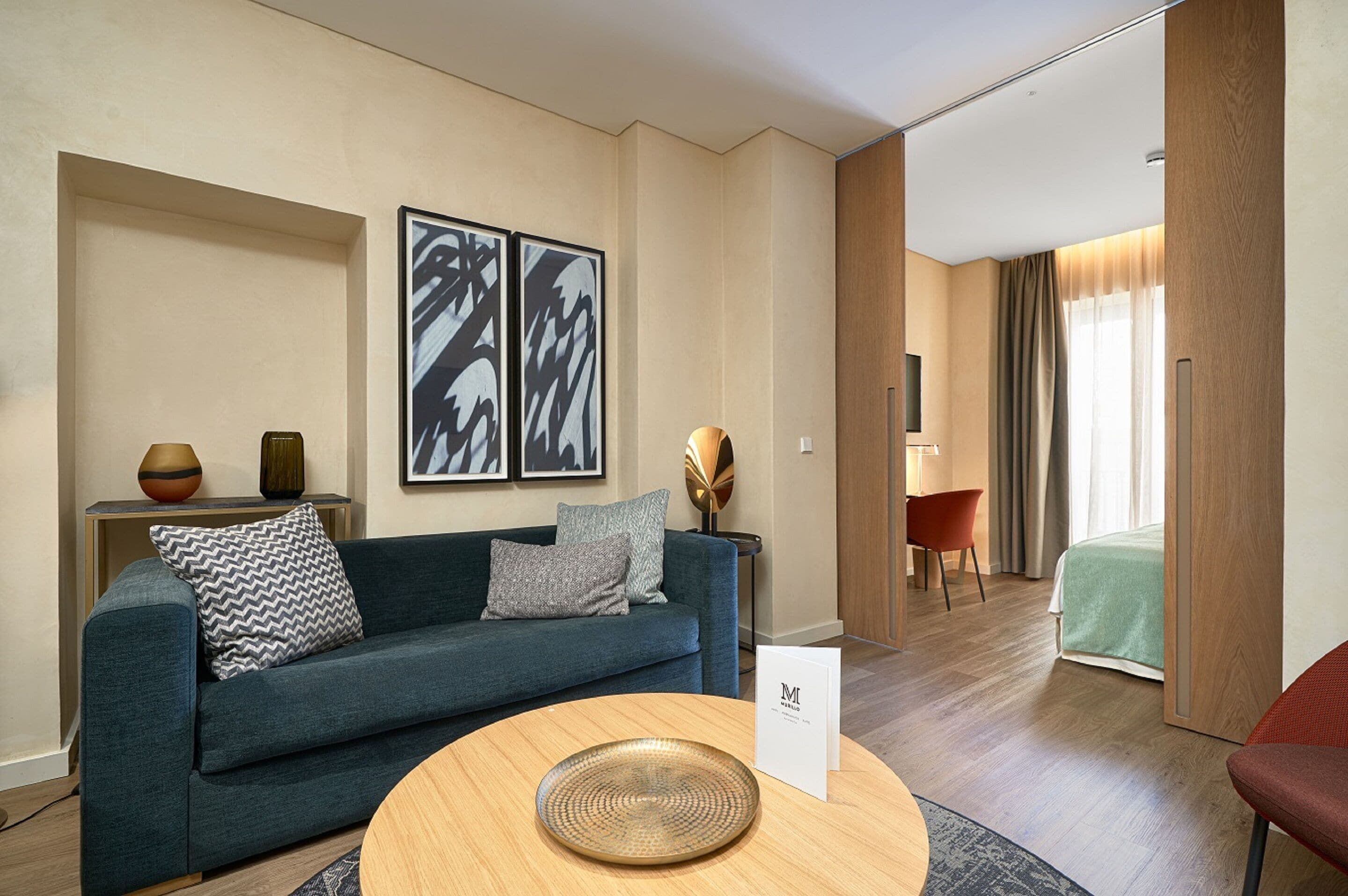 Et moderne hotelværelse med en sofa, et rundt træbord og en seng i baggrunden.