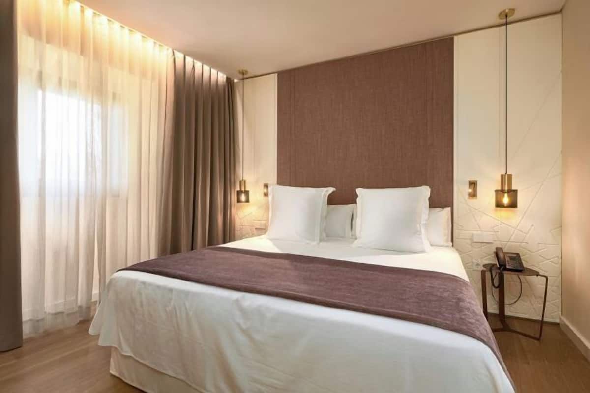 Ein Hotelzimmer mit einem großen Bett, Vorhängen, einem Nachttisch mit Lampe und einem an der Wand montierten Telefon.
