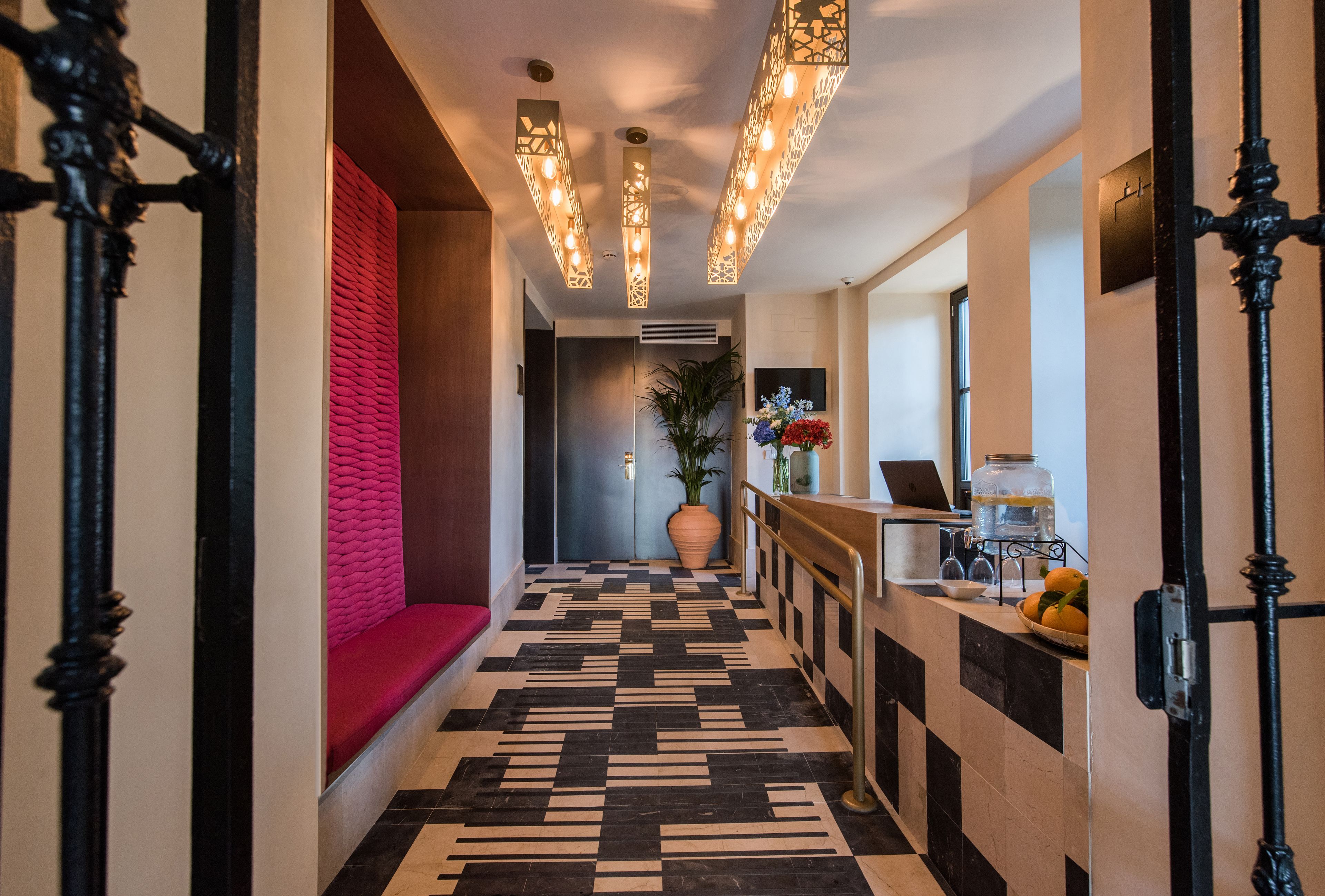 A hotel hallway with a checkered floor, a red bench, and a reception desk with a laptop and a vase of flowers.