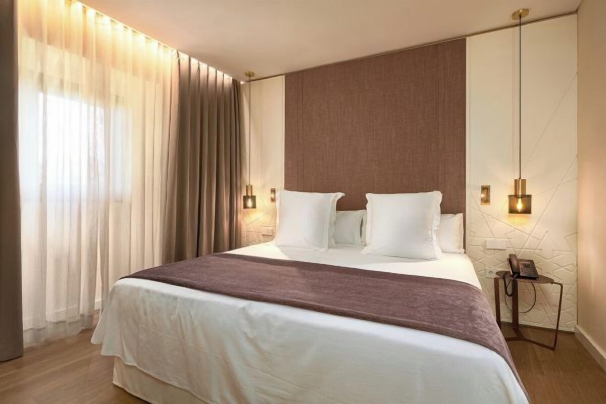 A hotel room with a large bed, curtains, a bedside table with a lamp, and a wall-mounted phone.
