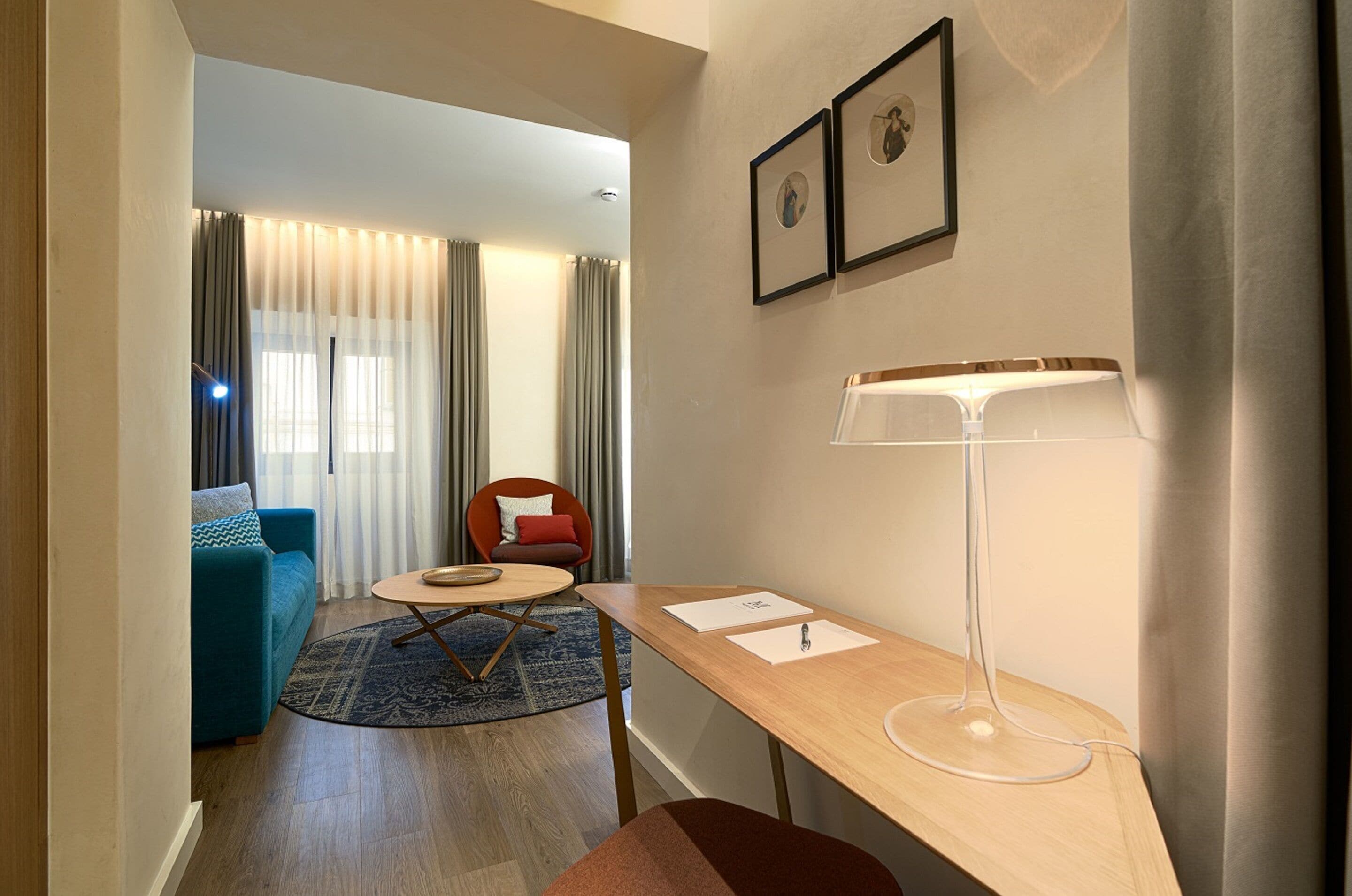 A modern living room with a sofa, a round table, a desk with a lamp, and a chair.