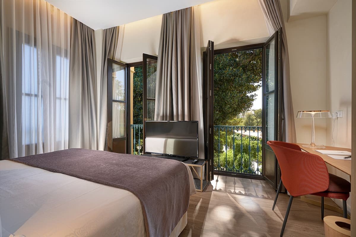 Ein Hotelzimmer mit einem großen Bett, einem Fernseher auf einem Schrank, einem Schreibtisch mit Stuhl und einem Balkon mit Blick auf Grünflächen.