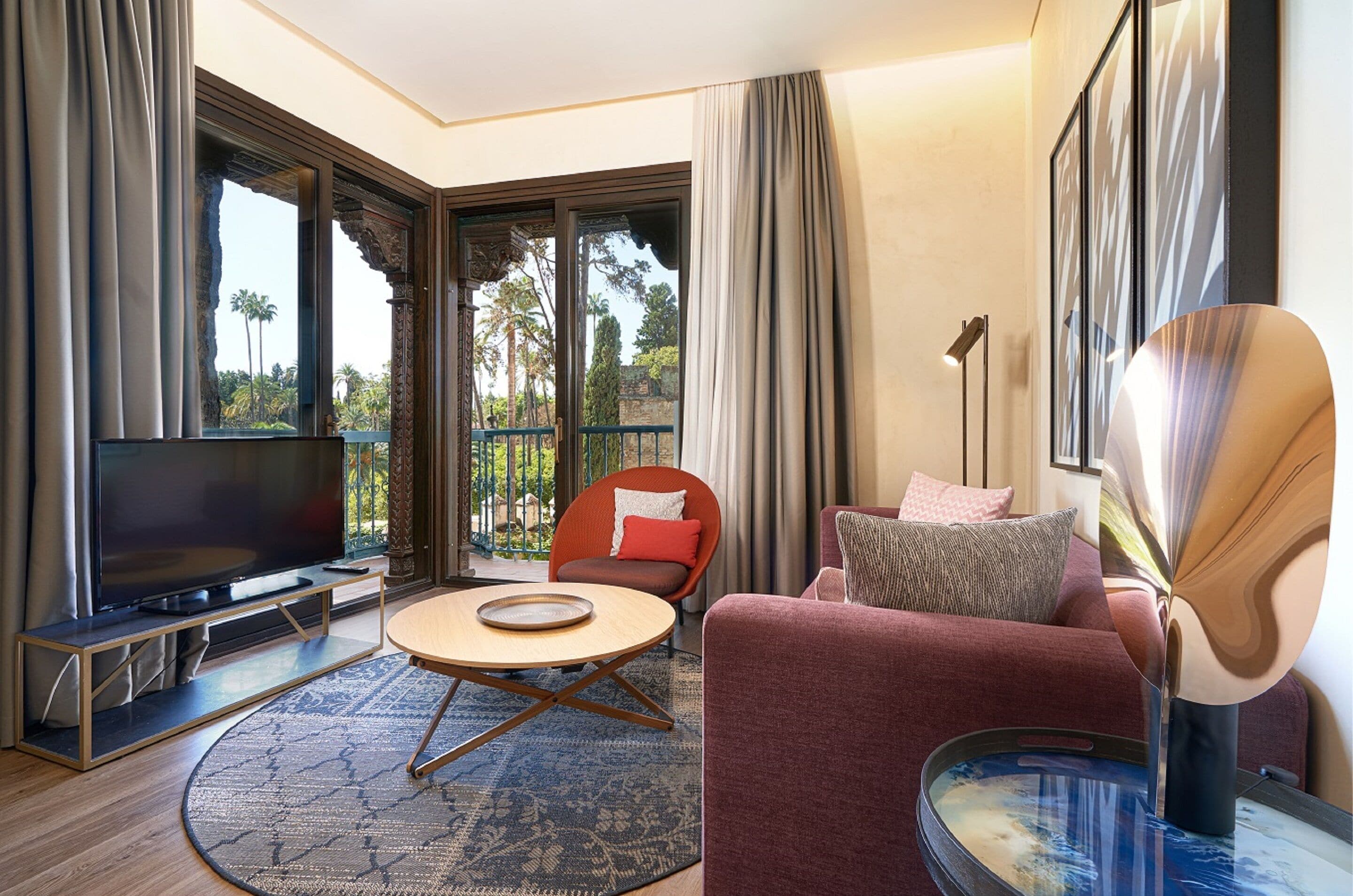 A modern living room with a flat-screen TV, a round coffee table, a red sofa, and a balcony with a view of palm trees.