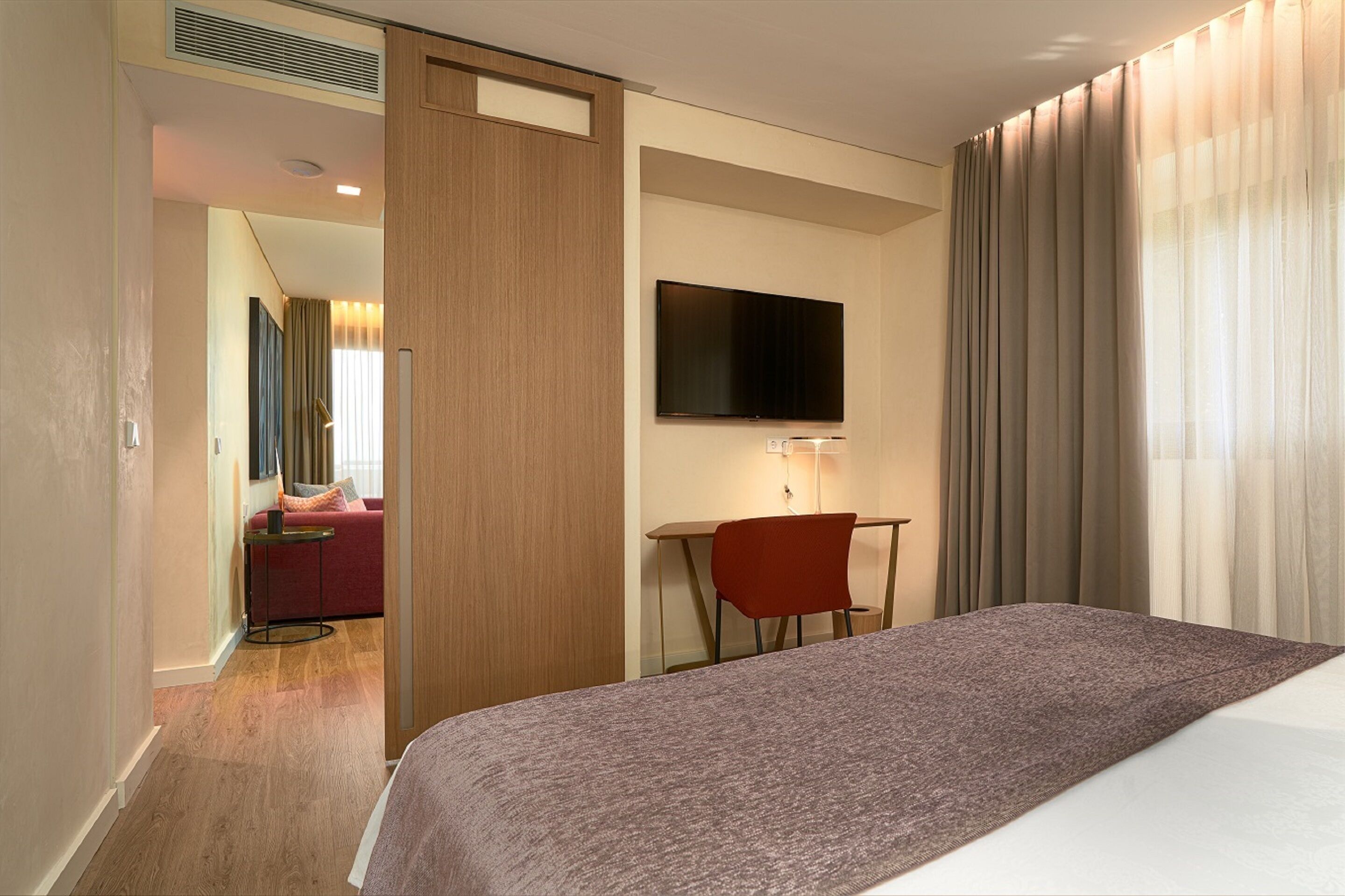 Une chambre d’hôtel comprenant un lit, un bureau avec une chaise, un téléviseur fixé au mur et une vue sur un canapé rouge situé dans la pièce adjacente.