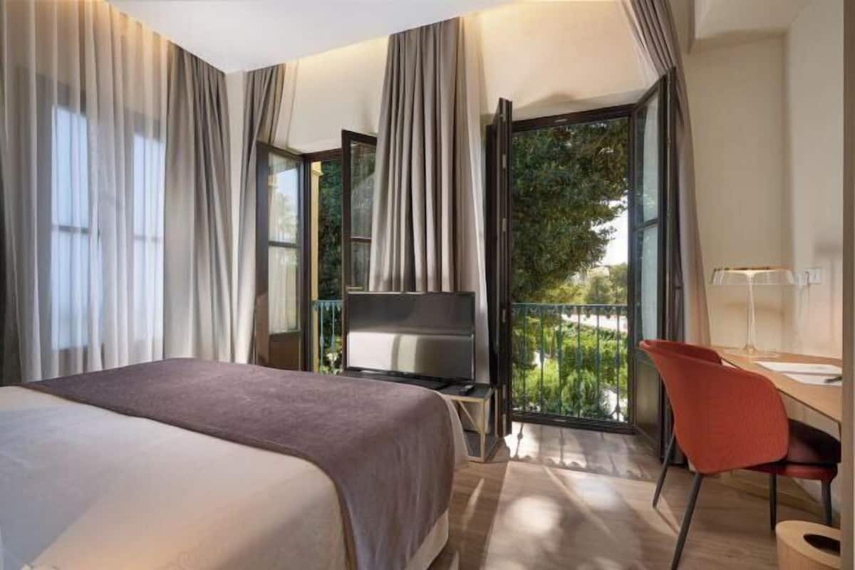 Ein Hotelzimmer mit Bett, Fernseher, Schreibtisch mit Stuhl und einem Balkon mit Blick auf Grünflächen.