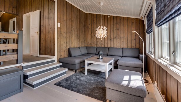 A modern living room with a grey sectional sofa, a white coffee table, and a black rug. There is a staircase leading to another room, and a large window with blinds on the right. - Bitigrenda H (Øystre Slidre)