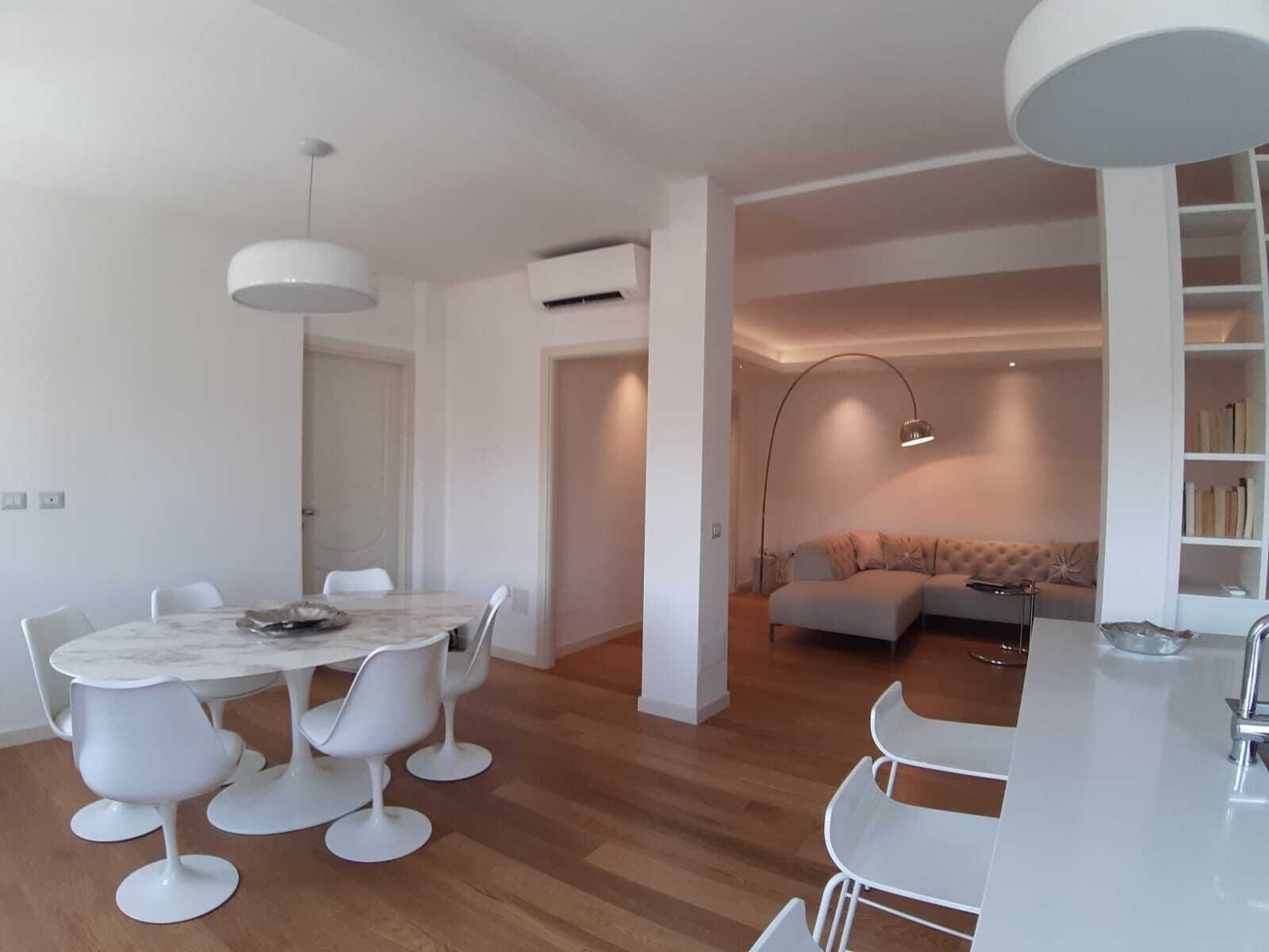 A modern living room with a round dining table, white chairs, a sofa, and a bookshelf.