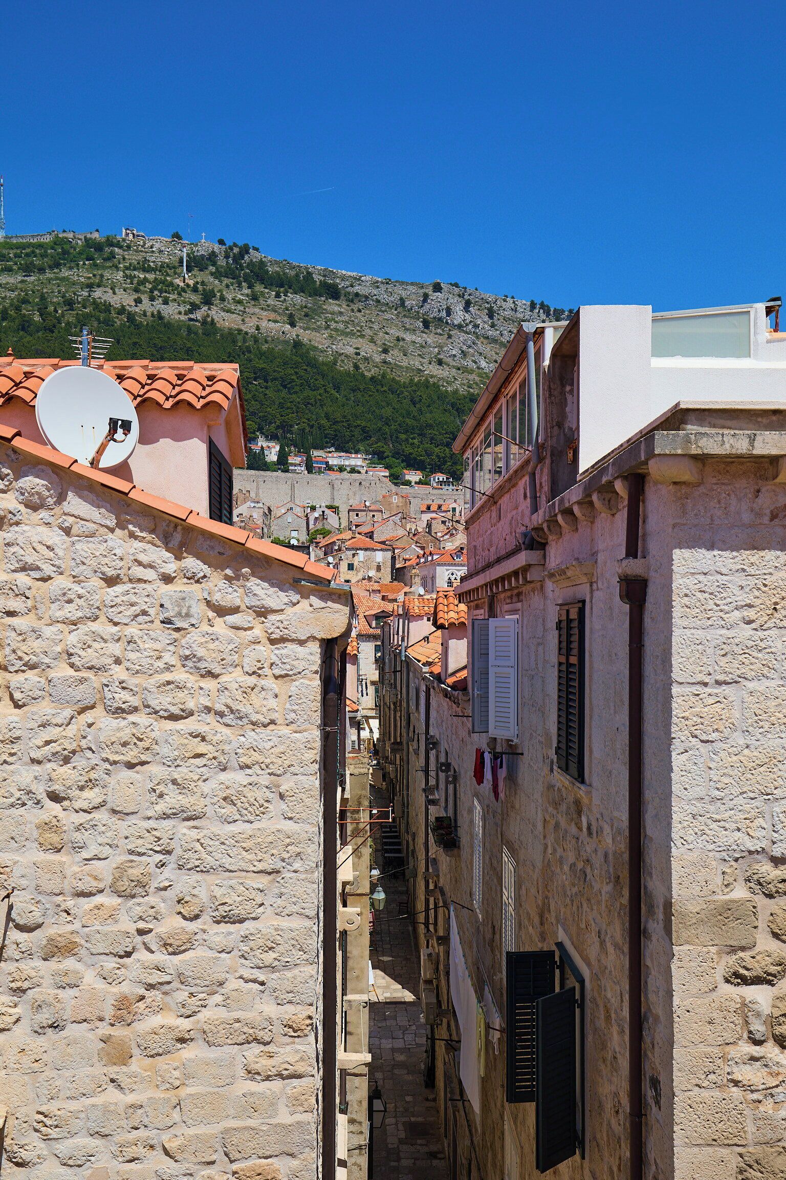 A narrow street with stone buildings, a satellite dish, and a mountainous backdrop.