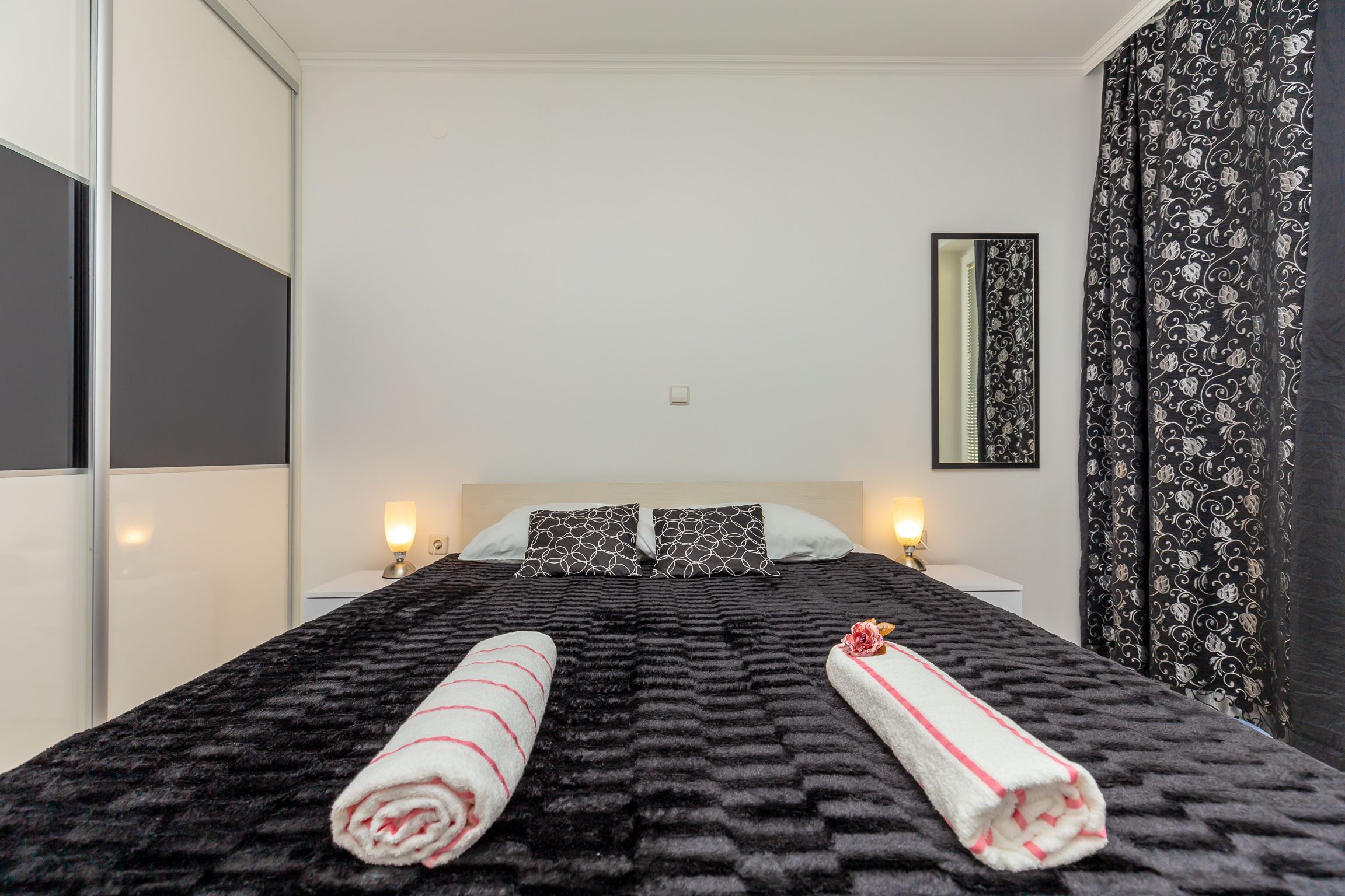 Una cama bien tendida con dos almohadas, un edredón negro con textura y dos toallas enrolladas con rayas rosas.