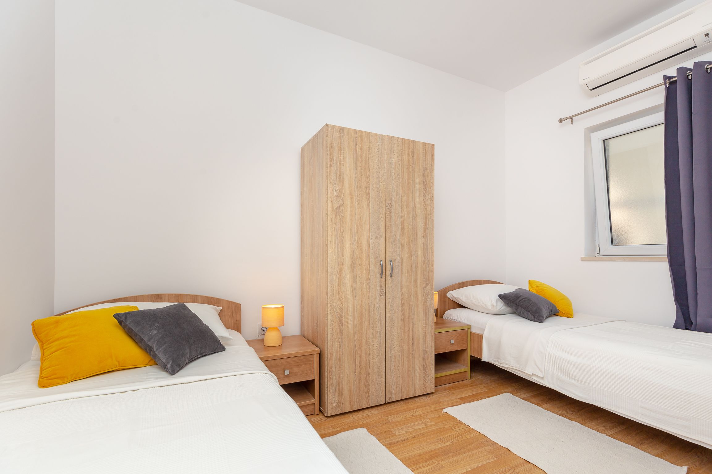 Un dormitorio con dos camas, un armario, una mesita de noche con lámpara y una ventana con cortinas.