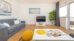 Uma sala de estar moderna com um sofá cinzento, uma TV de tela plana, uma mesa de centro branca com revistas e uma grande janela com vista.