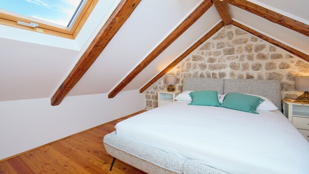 A bedroom with a sloped ceiling, stone wall, wooden beams, a bed, and bedside lamps. - Hedera Estate, Hedera A31 Apartment (Dubrovnik)