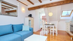 A living room with a blue sofa, a white table, a dining area with a table and chairs, a kitchen with an oven, and a skylight with a view of a building. - Hedera Estate, Hedera A31 Apartment (Dubrovnik)
