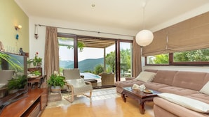 A spacious living room with a large sofa, a coffee table, and a TV. There is a dining area with a table and chairs, and a view of the outdoors. - Hedera Estate, House Hedera (Dubrovnik)