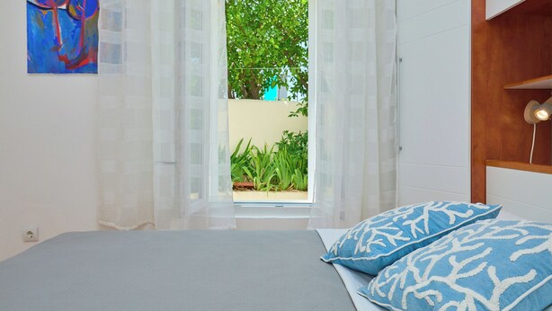 Quarto com cama, janela com cortinas leves e vista para o verde lá fora, além de um quadro na parede.