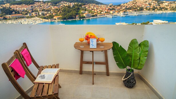 A balcony with a wooden table, a chair, a potted plant, and a view of a coastal town. - Skyscraper Apartment (Dubrovnik)