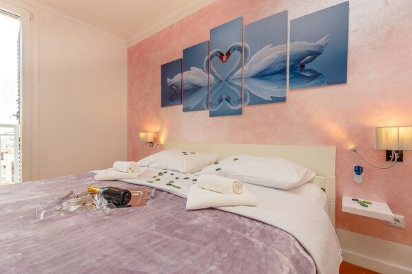 A hotel room with a bed, pillows, a bedside table, a wall-mounted artwork of swans, and a window with curtains.