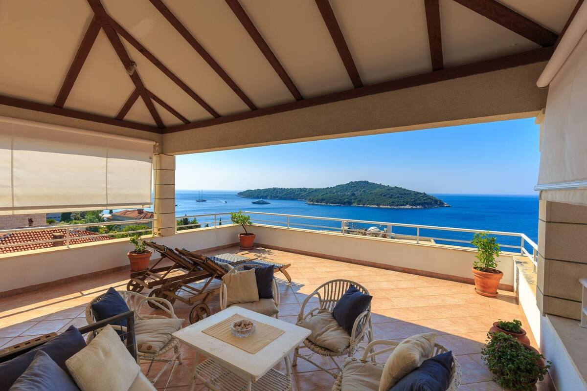 A covered outdoor seating area with wicker furniture, potted plants, and a view of the sea and a distant island.