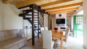 A modern living room with a wooden ceiling, a sofa, a dining table with chairs, a staircase, and a kitchen area with a TV and green door.
