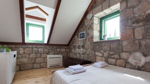 A bedroom with a stone wall, wooden beams, a bed with white linens, a white wardrobe, and a window with a green frame.