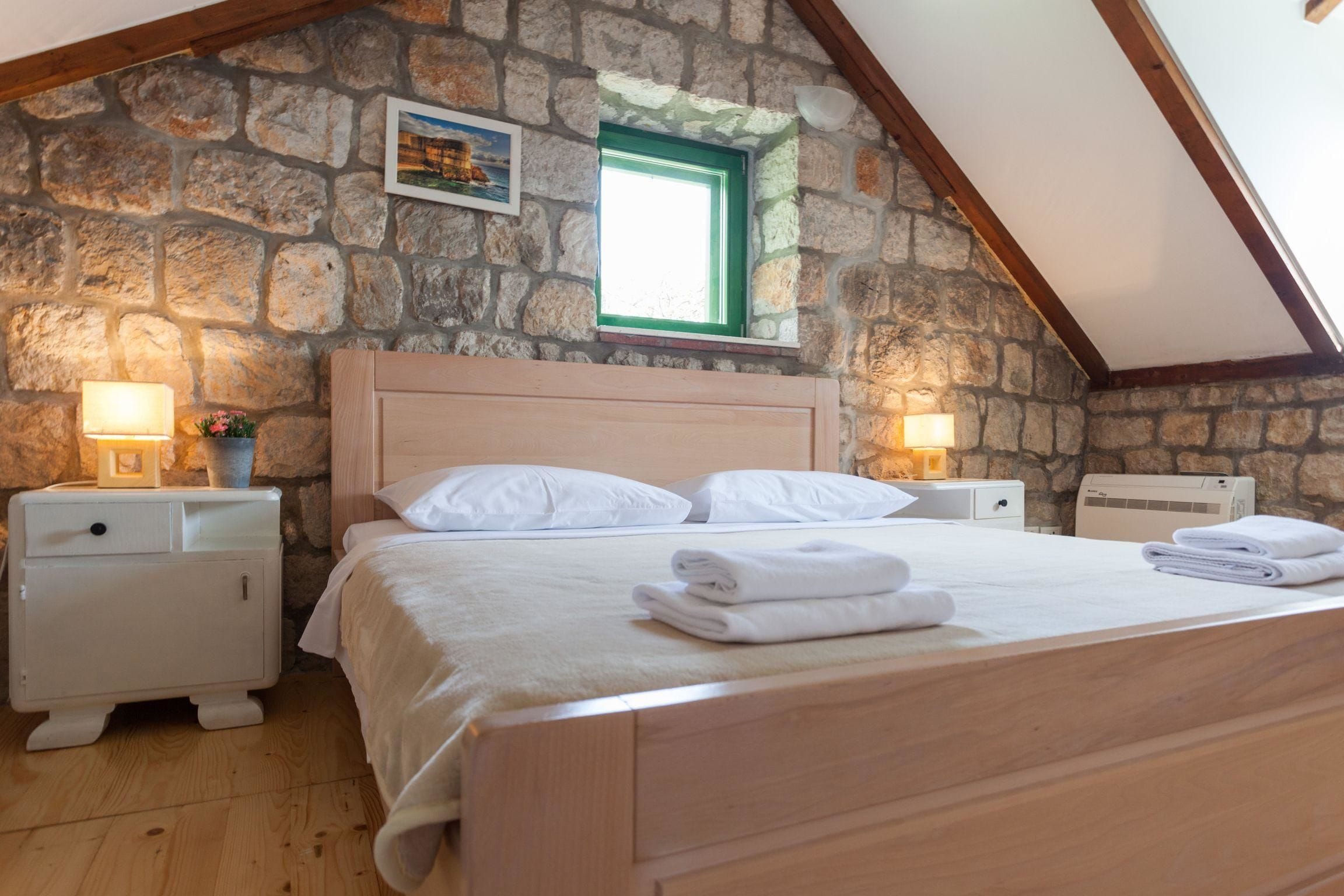 Un dormitorio con pared de piedra, techo de madera, cama con ropa blanca, dos mesitas de noche con lámparas y un ventanal pequeño.