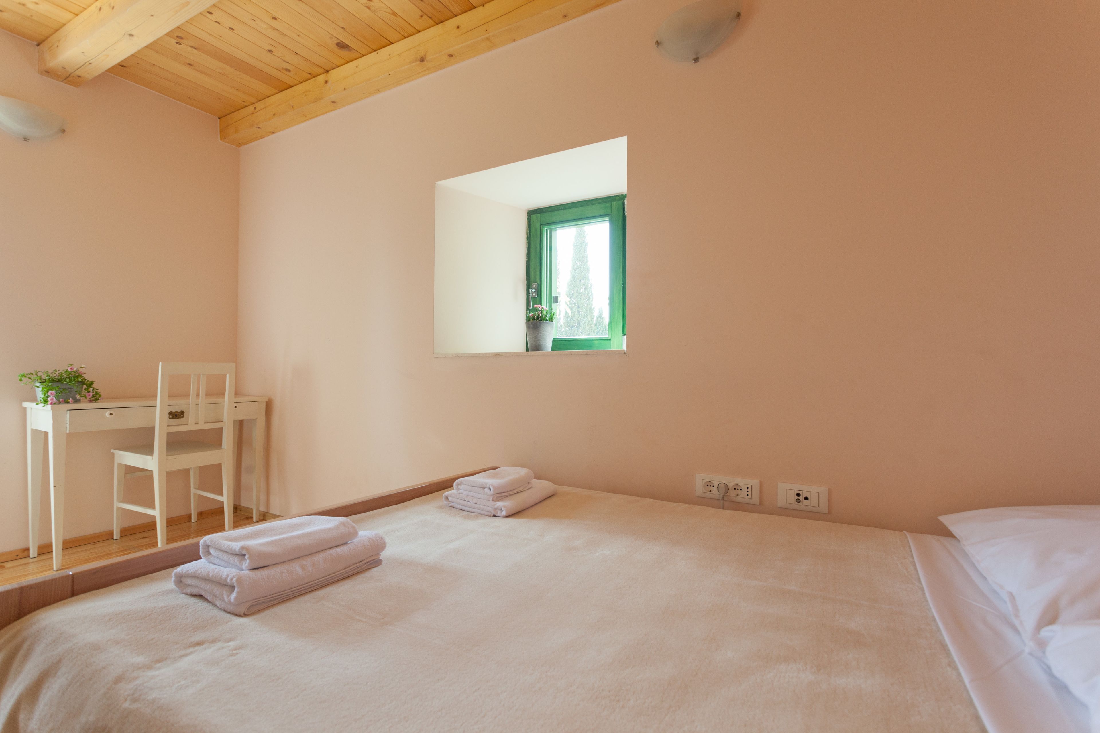 A bedroom with a bed, a desk, a chair, and a window with a view of greenery.