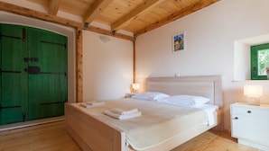 A bedroom with a wooden ceiling, a green door, a bed with white linens, a nightstand with a lamp, and a small window with a view of greenery.