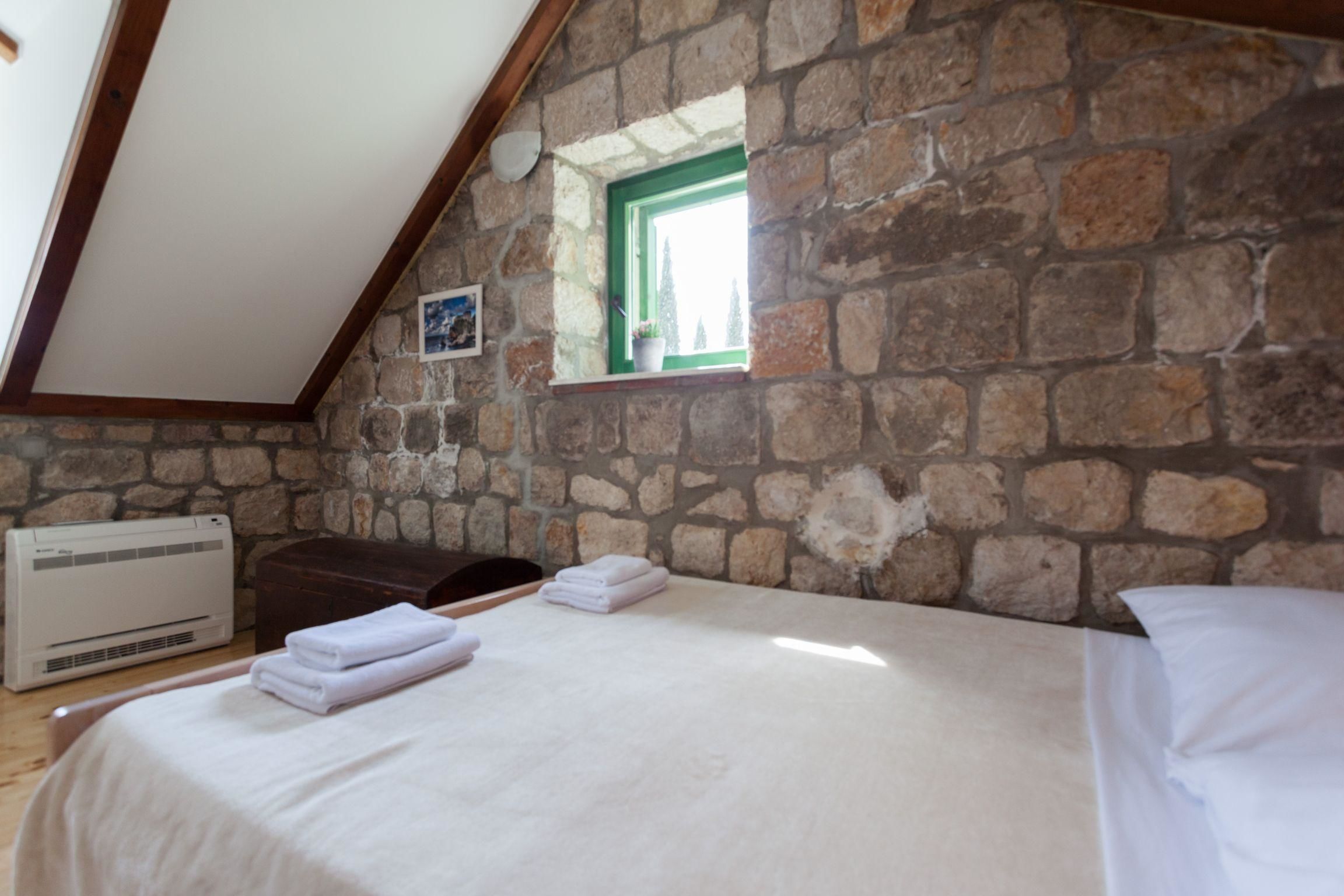 Une pièce avec un mur en pierre, une fenêtre à cadre vert, un lit avec du linge de lit blanc et un appareil de climatisation.