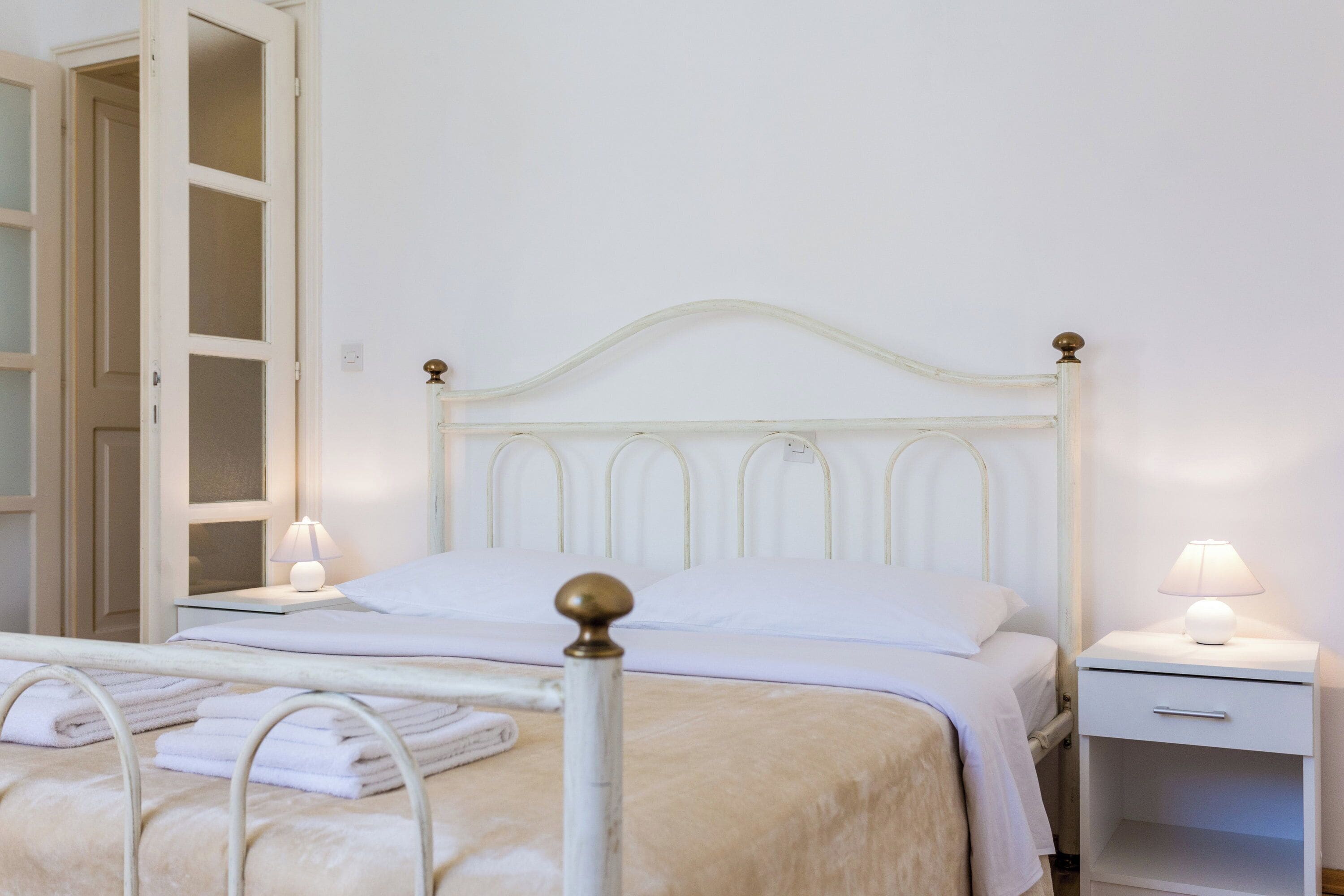 Un lit bien fait, avec du linge de lit blanc, une table de chevet avec une lampe, et une porte qui donne sur une autre pièce.