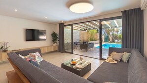 A modern living room with a large grey sofa, a flat-screen TV, and a glass door leading to an outdoor seating area with a pool. - Hedera Estate, Villa Hedera II (Dubrovnik)