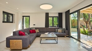 Un salon moderne avec un canapé d’angle gris, une table basse en bois et de grandes fenêtres donnant sur un paysage verdoyant.