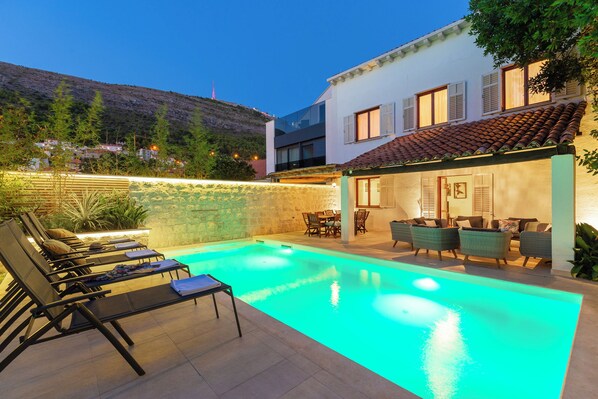 A swimming pool with lounge chairs and a dining area, surrounded by a stone wall and a hillside view. - Hedera Estate, Villa Hedera (Dubrovnik)