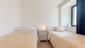 Une petite chambre propre, avec deux lits simples, une table de chevet avec une lampe et une fenêtre avec des rideaux.