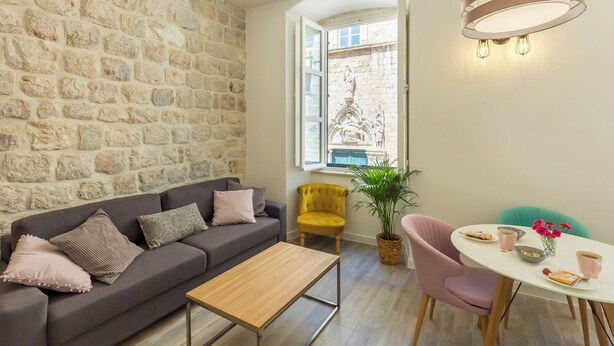A cozy living area with a stone wall, a sofa, a coffee table, a dining table with chairs, and a window with a view of a building. - Hedera Estate, Hedera A2 Apartment (Dubrovnik)