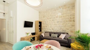 A modern living room with a stone wall, a grey sofa, a wooden cabinet, and a round table with a vase of flowers. - Hedera Estate, Hedera A2 Apartment (Dubrovnik)