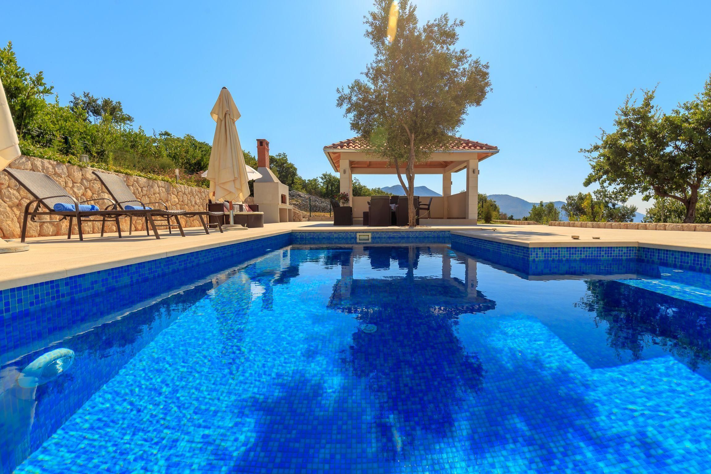 Une piscine dotée de chaises longues et d’un kiosque, entourée d’arbres et d’un mur de pierre.