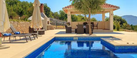 Una piscina con sillones de descanso y un pabellón, rodeada de follaje y un muro de piedra.