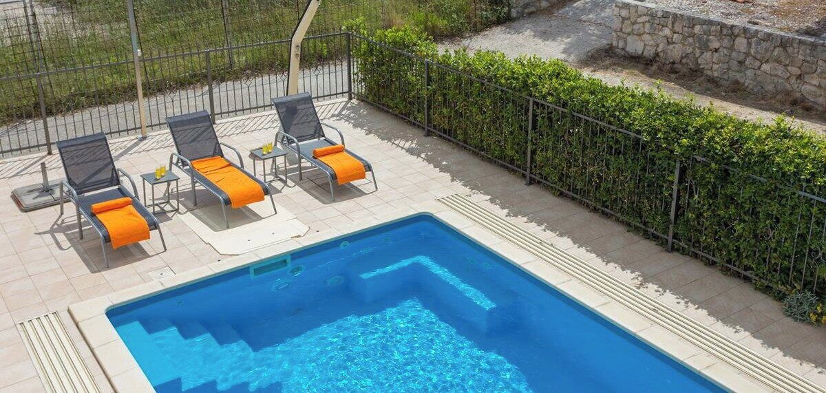 Une piscine rectangulaire, bordée d’un revêtement de carrelage, entourée d’une clôture en métal et d’une haie.