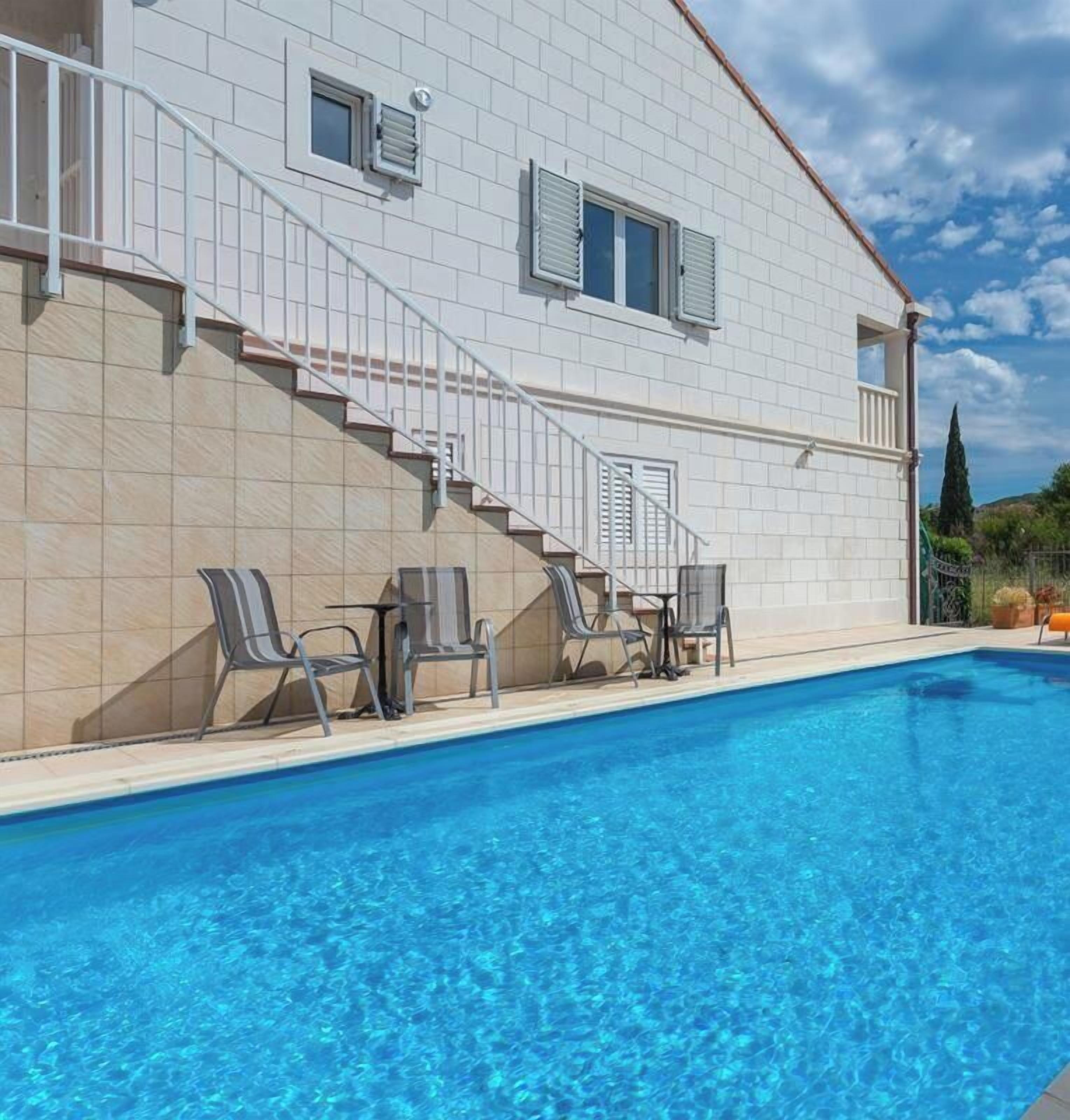 Ein Schwimmbecken mit einem weiß gefliesten Gebäude im Hintergrund, einer Treppe und Außenmöbeln.