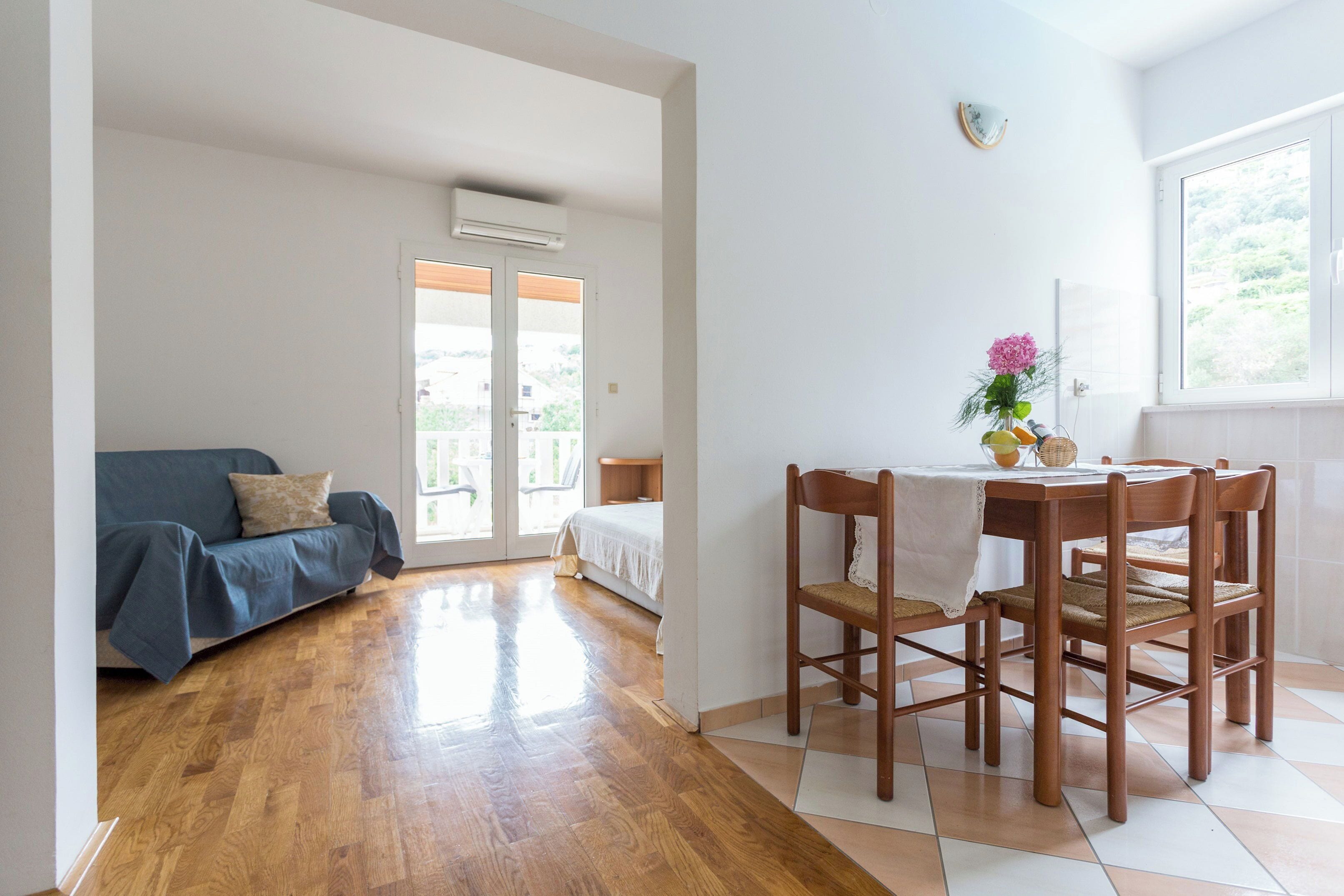 A room with a wooden floor, a blue sofa, a dining table with chairs, and a window with a view.
