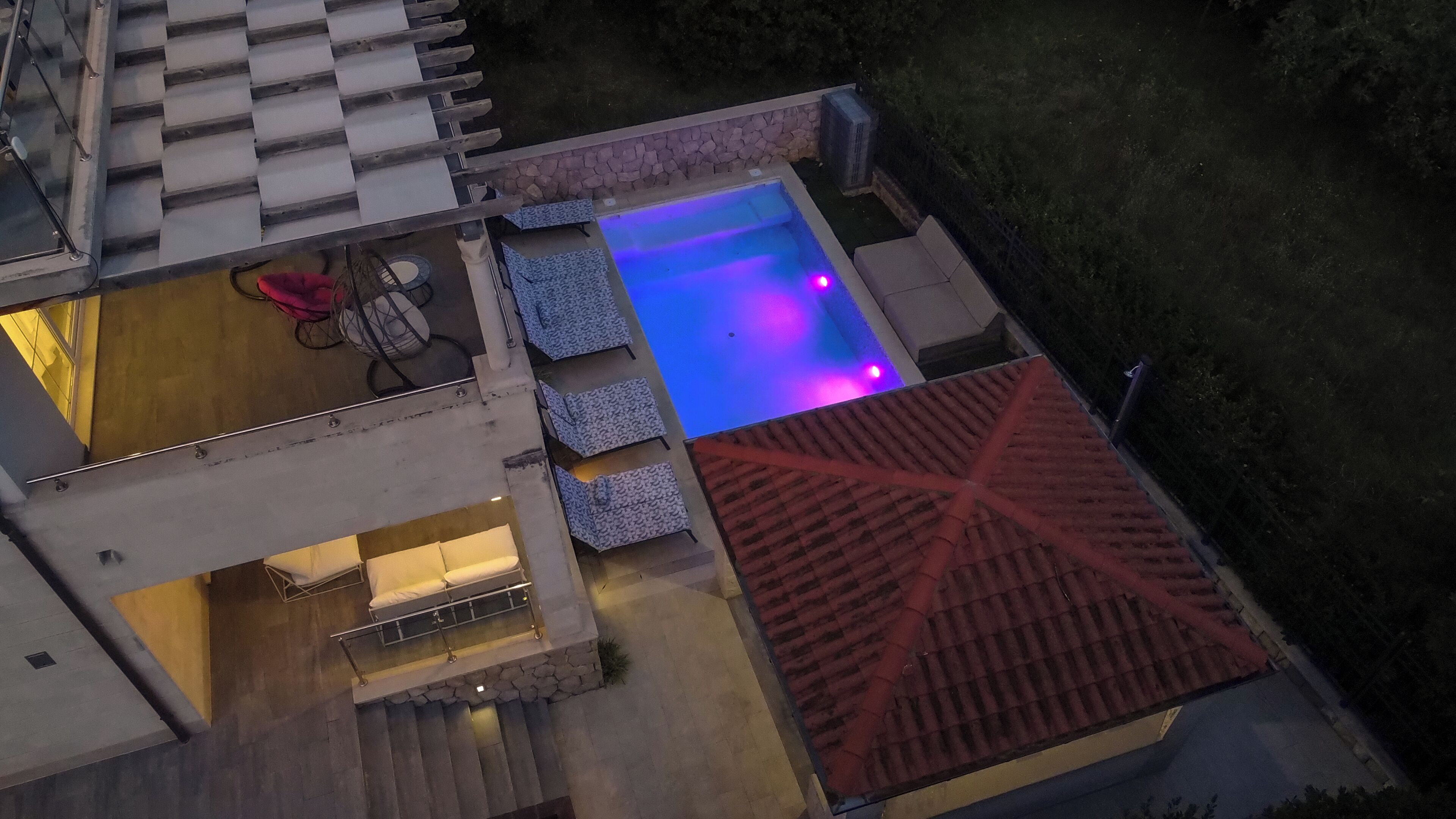 A rooftop pool area with a blue-lit pool, outdoor seating, and a red-tiled roof.