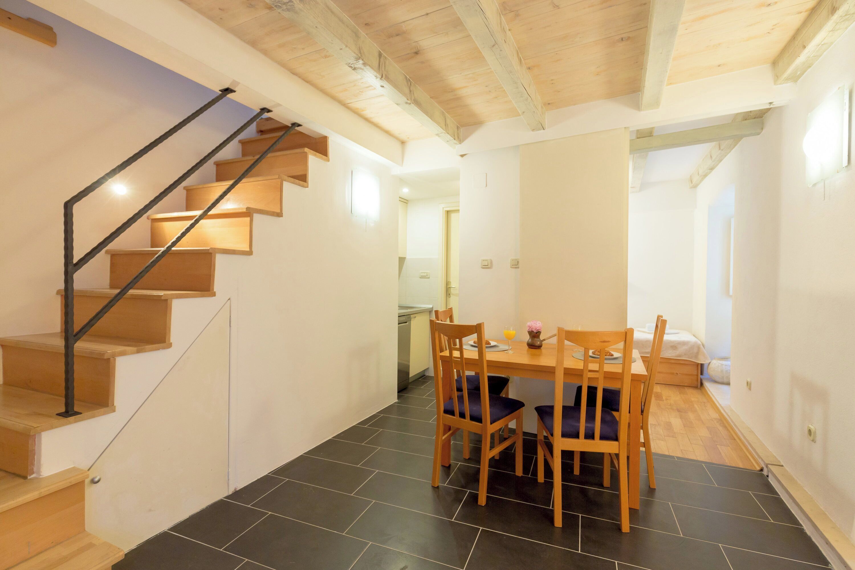 Una escalera con escalones de madera y barandillas de metal, un comedor con una mesa y sillas, y una cocina al fondo.
