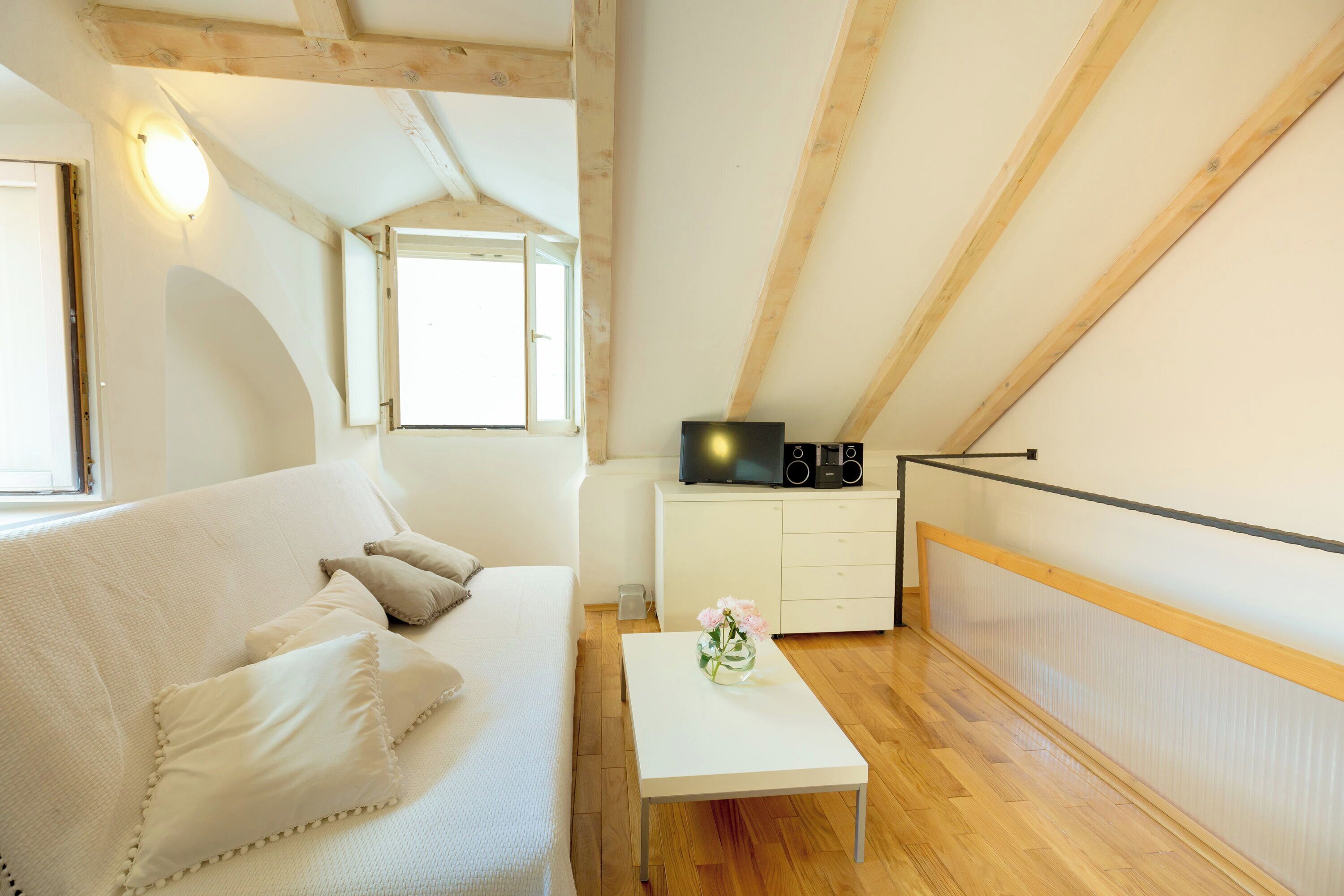 Une pièce avec un canapé blanc, une petite table blanche ornée d’un vase, un plancher en bois, un meuble blanc sur lequel se trouve un téléviseur, et une fenêtre donnant sur un paysage.