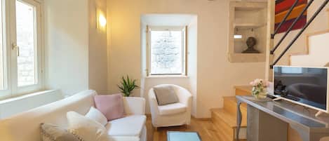 Una sala de estar con un sofá, una silla, un escritorio con una computadora, una escalera y un ventanal con vista a un muro de piedra.