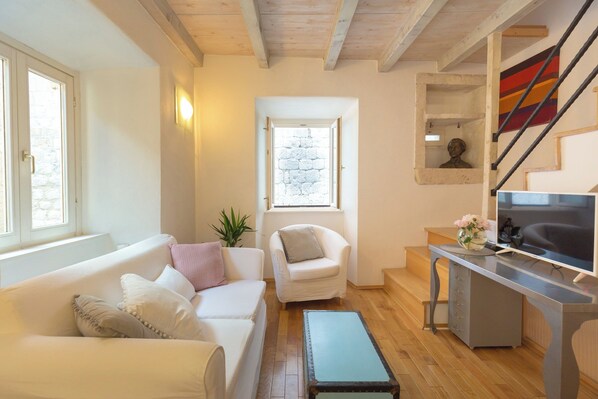 A living room with a sofa, a chair, a desk with a computer, a staircase, and a window with a view of a stone wall.