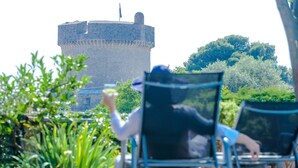 A person sitting on a chair facing a castle-like structure, with greenery and a clear sky in the background. - Villa Donna Paola (Dubrovnik)